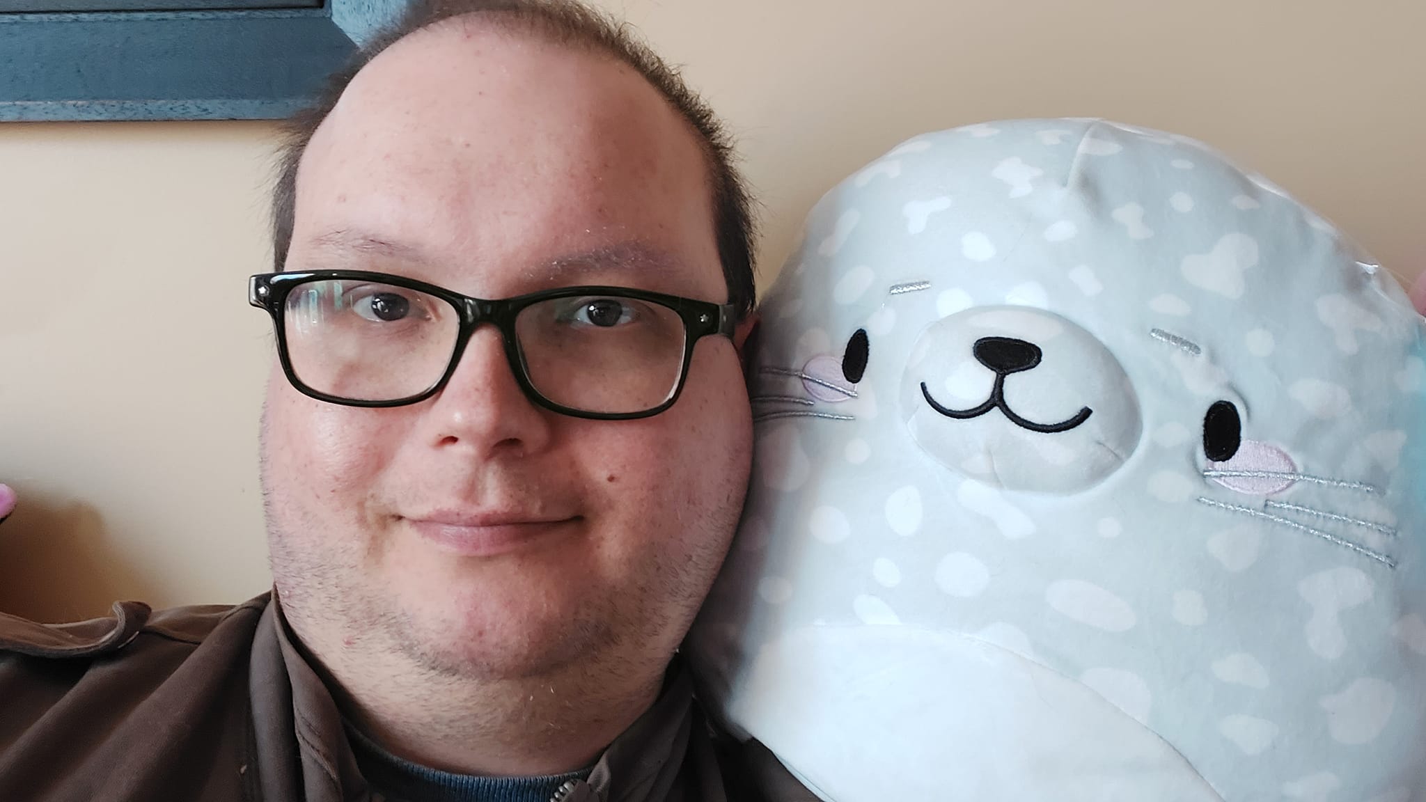 A man sits on a couch, a seal Squishmallow rests on his sholder and takes up half the frame